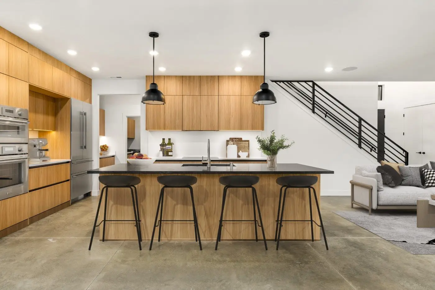 Modern kitchen with wood cabinets, black countertop island with four black barstools, stainless steel appliances, pendant lights, and an open living area with stairs in the background.