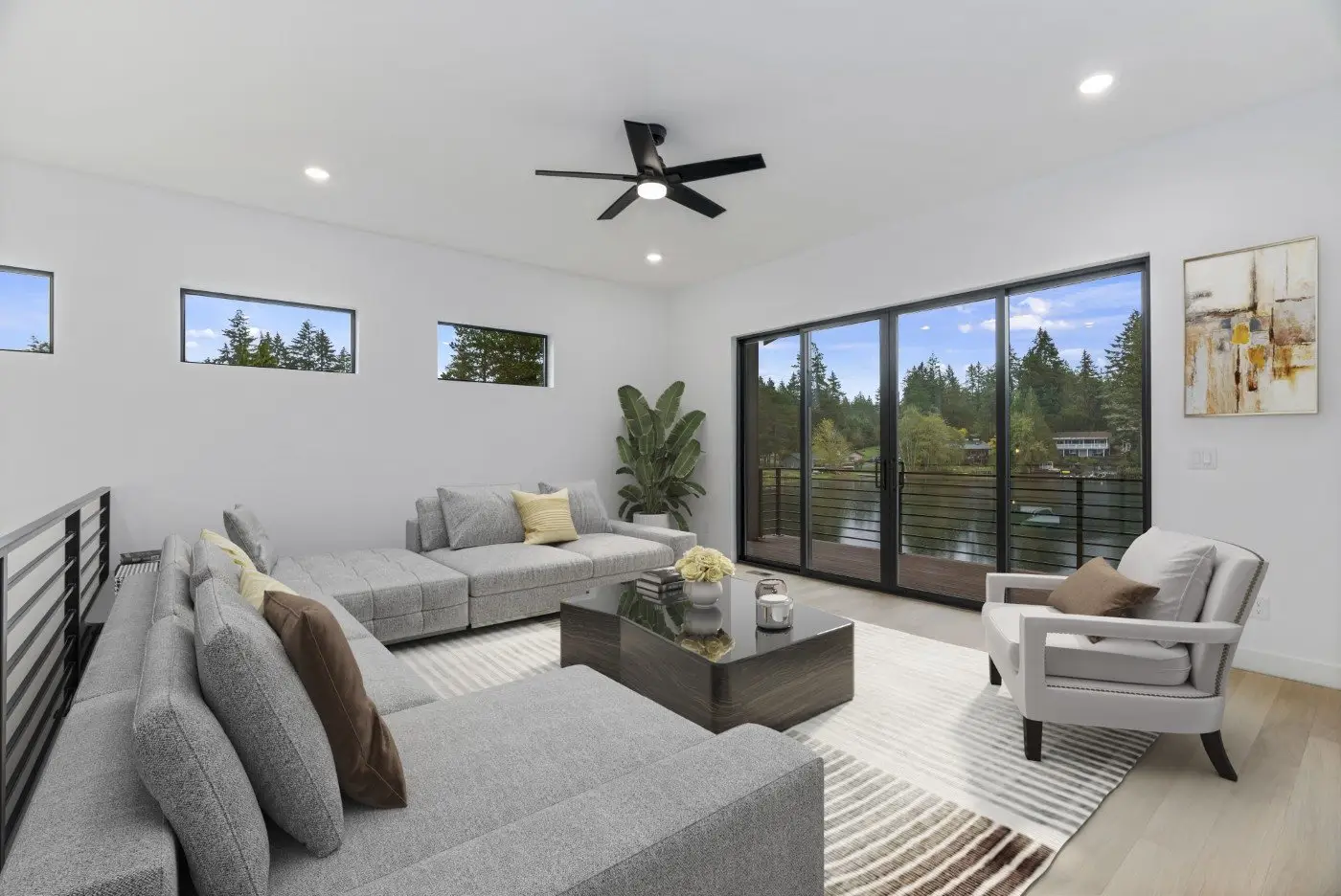 Modern living room with large gray sectional sofa, white armchair, glass coffee table, and plant. Floor-to-ceiling windows show greenery outside. Room features neutral tones and wall art.