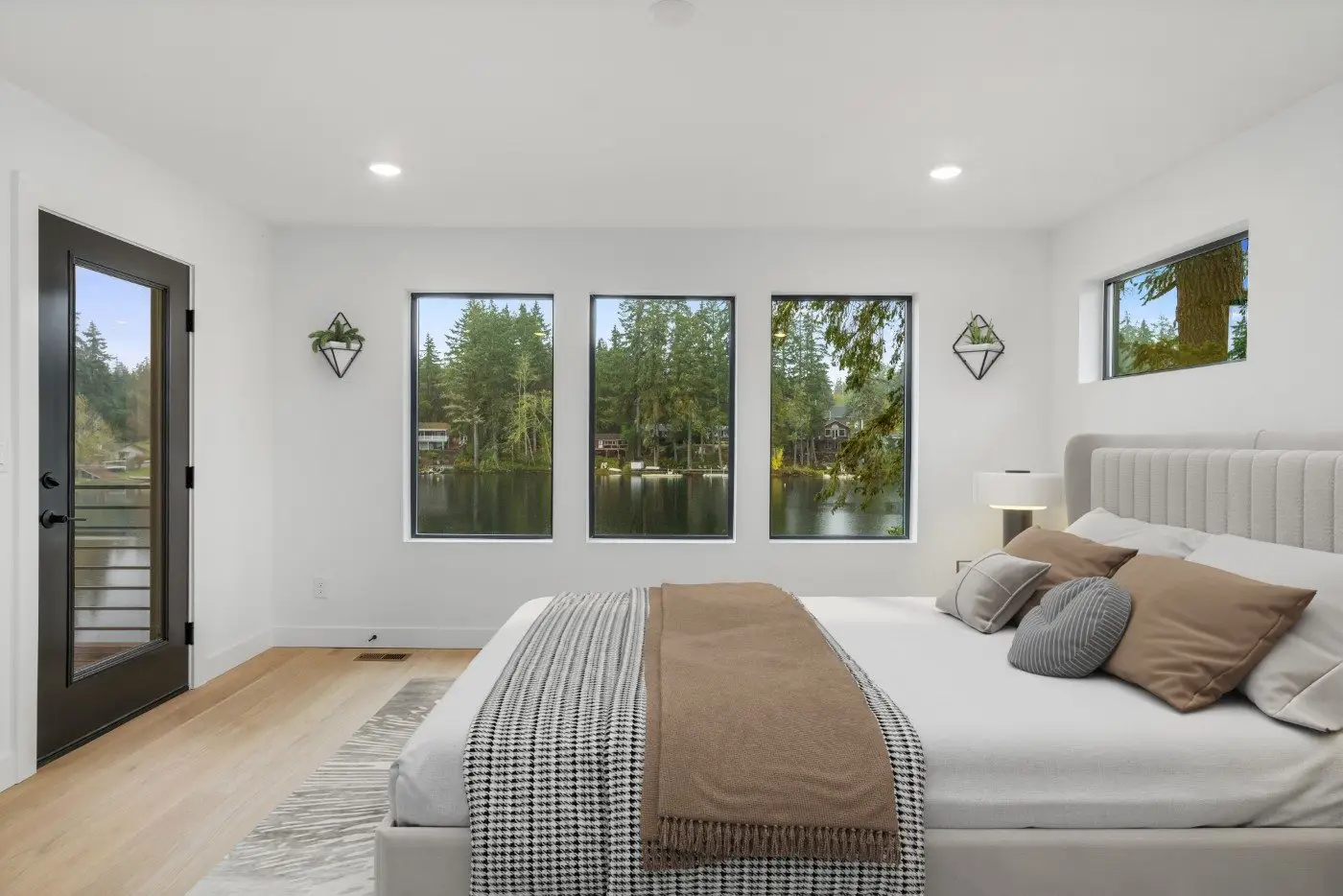 A modern bedroom with large windows showing a lake and trees outside. The room has a neatly made bed with neutral-colored bedding, a light wood floor, and a black-framed glass door leading to a balcony.