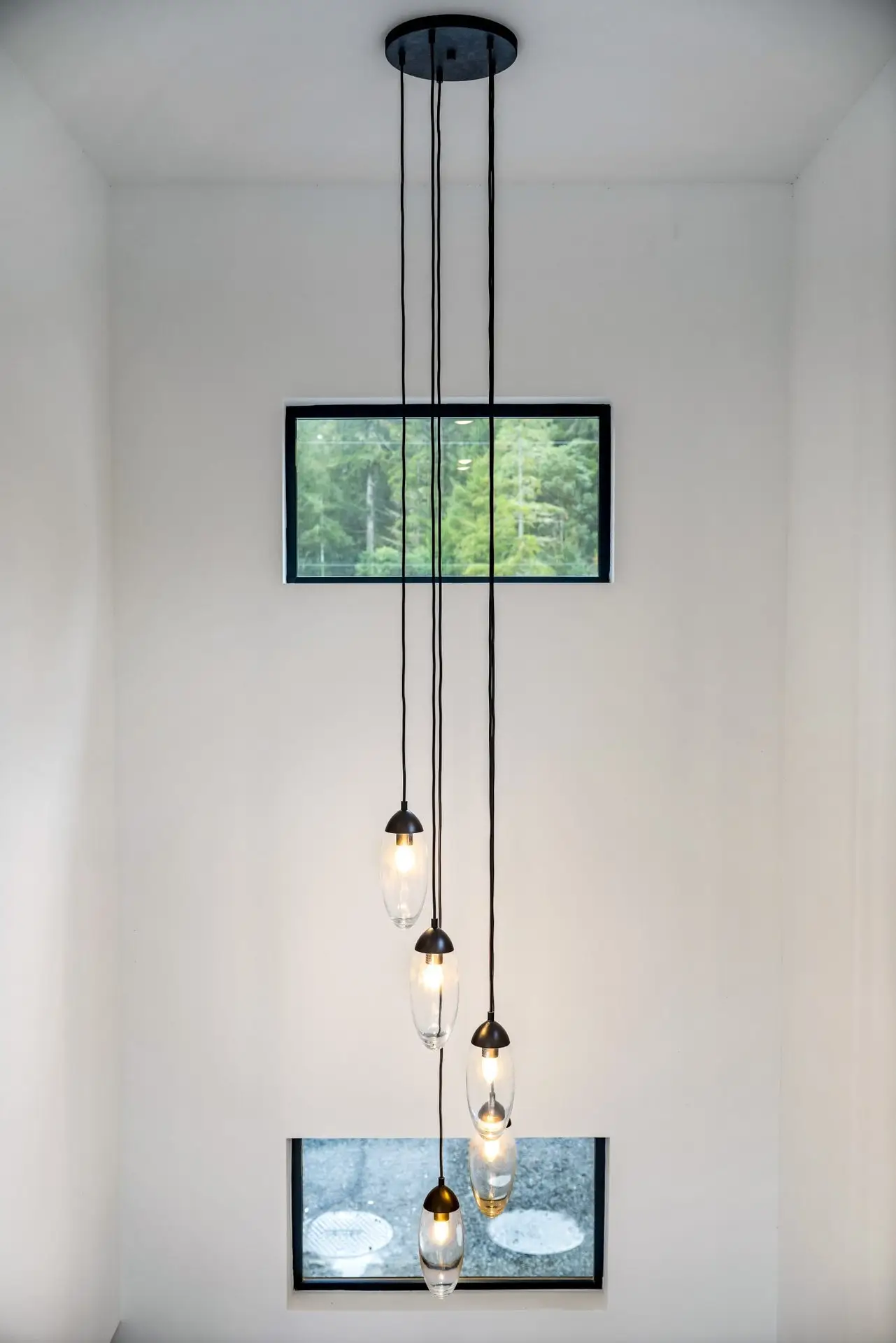A modern pendant light with five hanging glass bulbs descends from a ceiling in front of two rectangular windows, with green trees visible outside the top window.