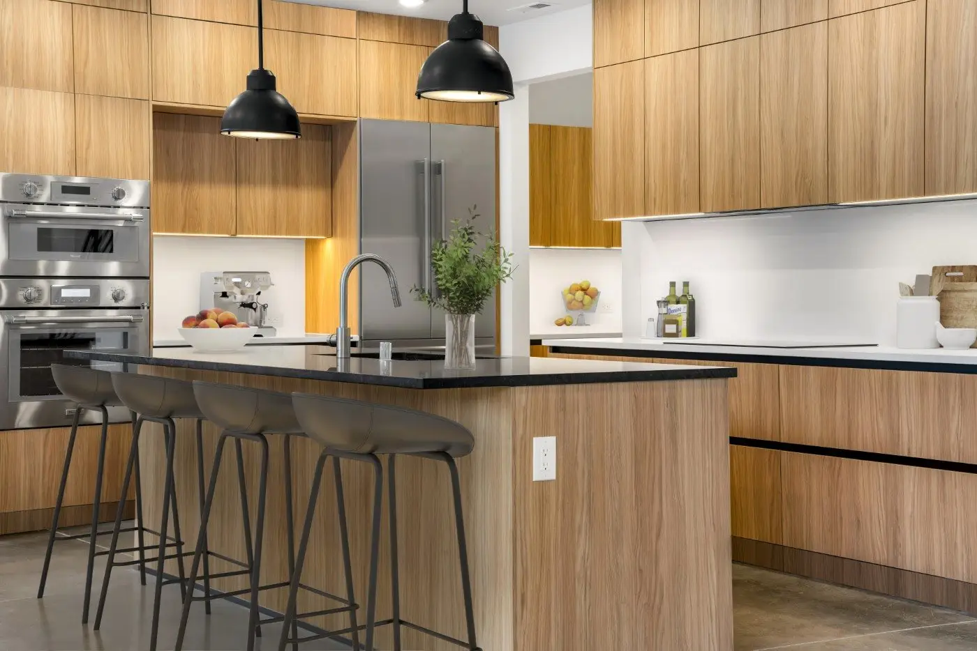 Modern kitchen with wood cabinets, black countertops, an island with four bar stools, pendant lights, built-in stainless steel appliances, and a bowl of apples on the counter.