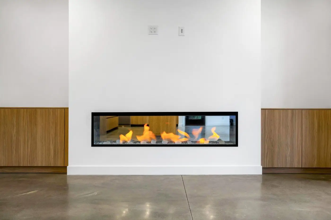 A modern built-in rectangular gas fireplace with visible flames is set in a white wall, featuring wooden paneling on both sides and a polished concrete floor.