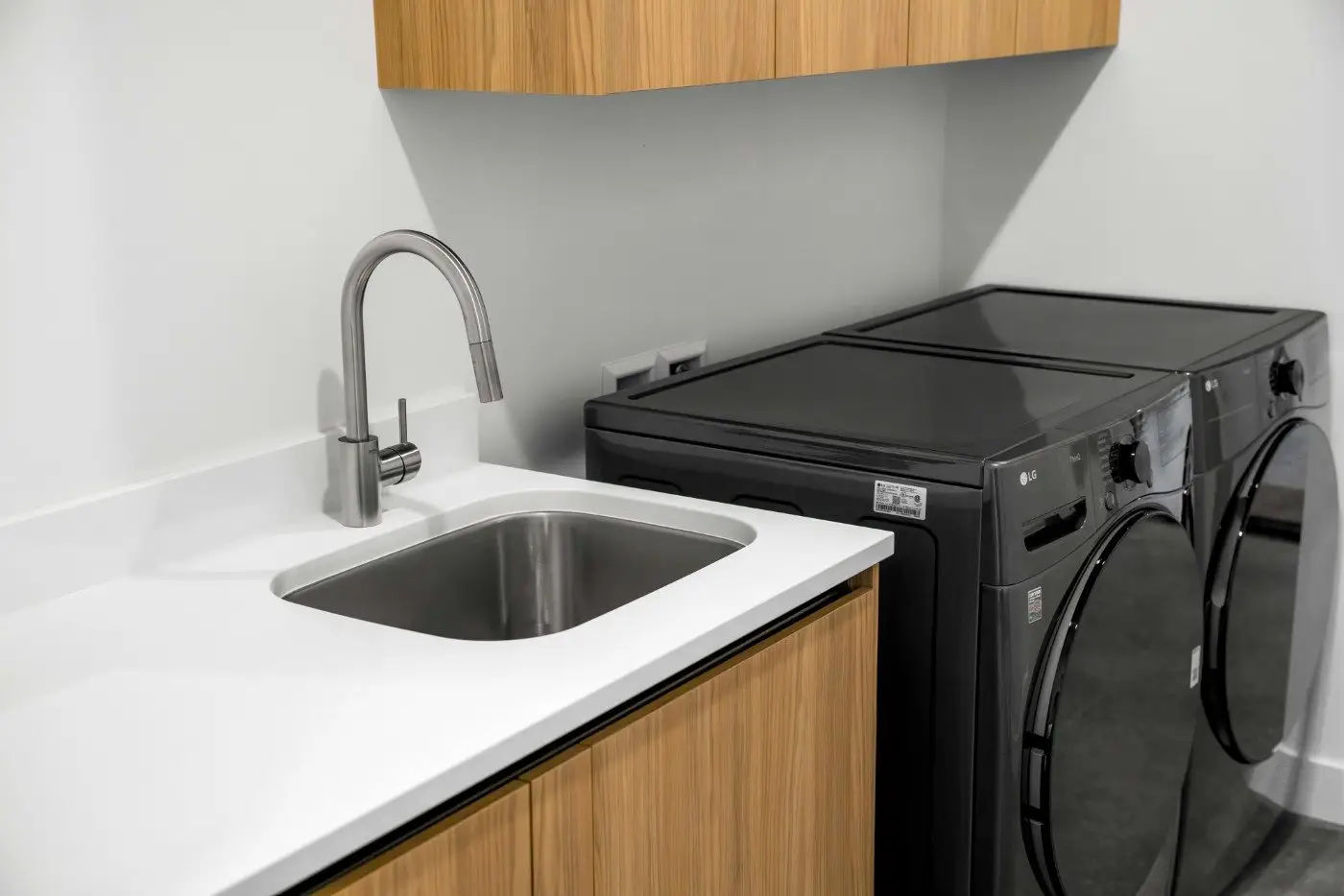 A modern laundry room with a stainless steel sink, wooden cabinets, and a black front-loading washer and dryer set against a light gray wall.