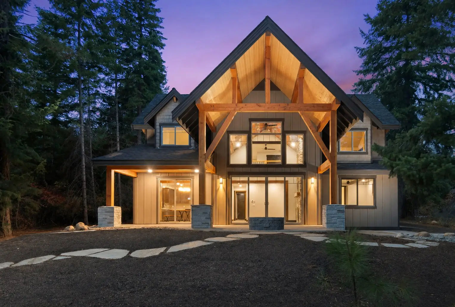 Timber-frame custom home glowing at dusk among tall evergreens, built by Doyle Construction