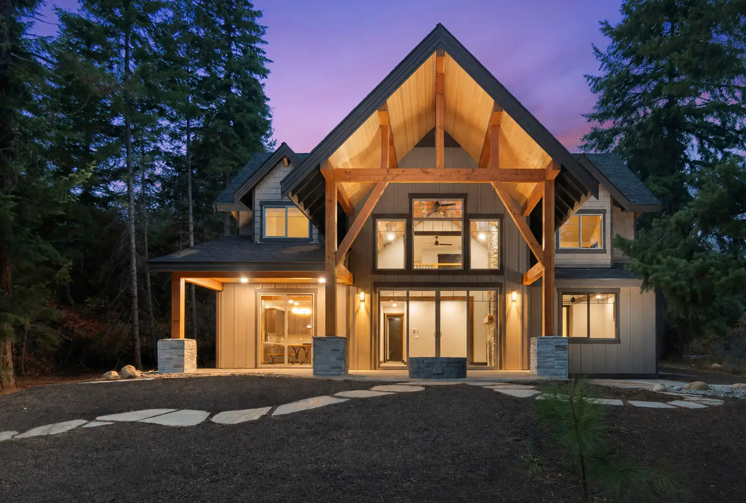 A custom timber-frame home built by Doyle Construction, glowing at dusk among tall evergreens