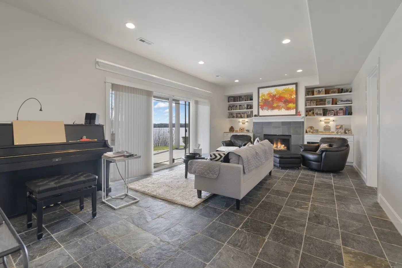A modern living room with gray tiled floor, gray sofa, two black armchairs, a fireplace with colorful art above it, built-in shelves, a piano, and large sliding glass doors offering a view outside.
