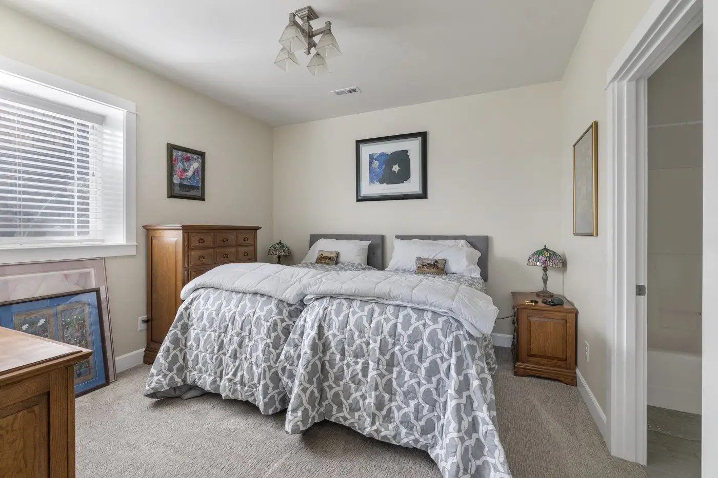 A cozy bedroom with two twin beds pushed together, gray patterned bedding, wooden dressers, framed art on the walls, and a window with blinds letting in natural light. A small lamp sits on a bedside table.