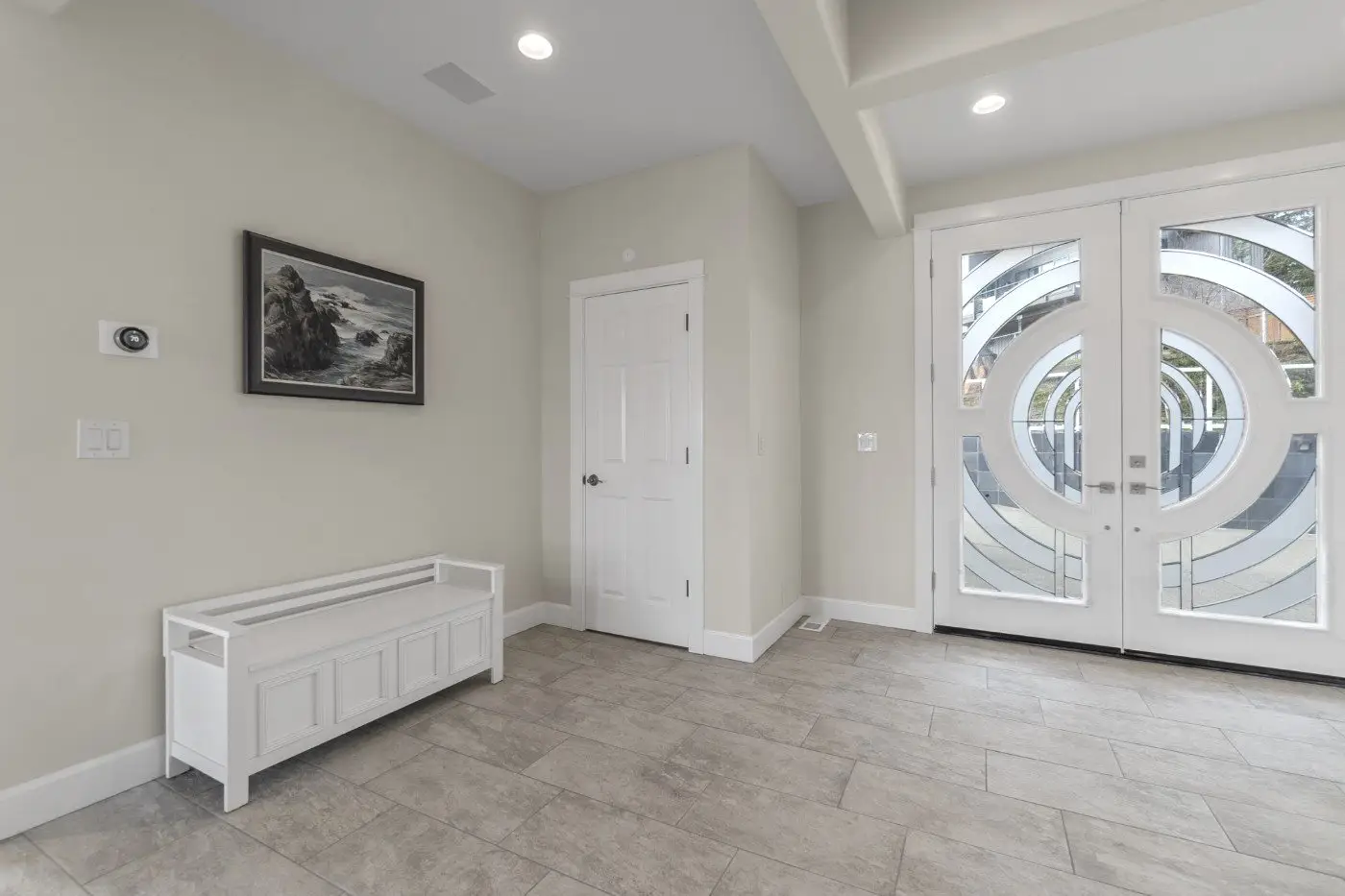A modern entryway with light tile flooring, double glass front doors featuring circular designs, a white bench with storage, a closed white door, and a framed landscape artwork on a beige wall.