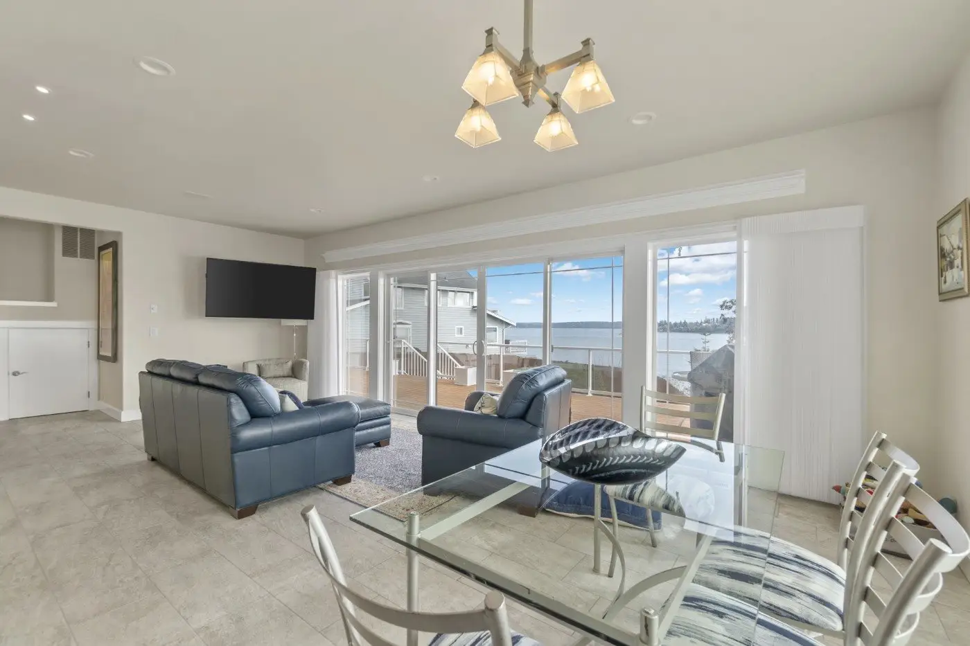 A bright, modern living and dining area with glass doors opening to a deck overlooking water and neighboring houses. Features a glass dining table, blue sofas, and a wall-mounted TV.