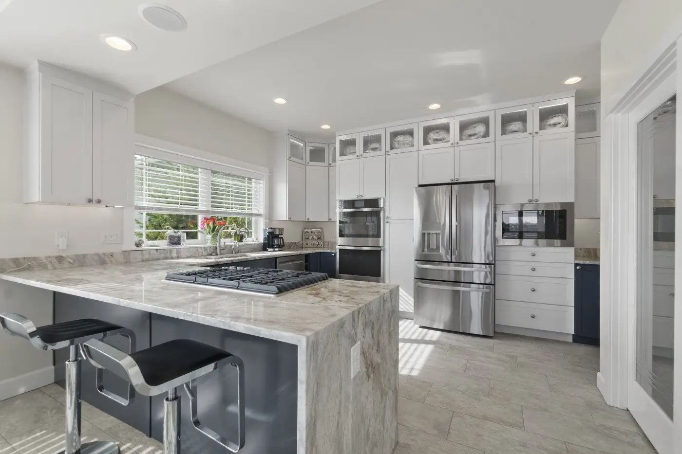 Modern kitchen with white cabinets, stainless steel appliances, marble countertops, a large island with a built-in stove, two barstools, and a window letting in natural light.