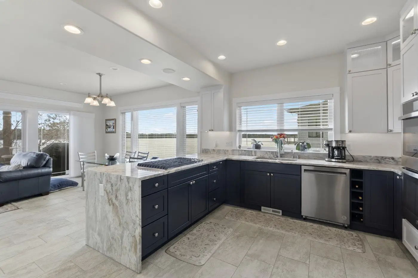 Modern kitchen with navy blue lower cabinets, white upper cabinets, marble countertops, stainless steel appliances, large windows, and an adjacent dining area with glass doors leading outside.