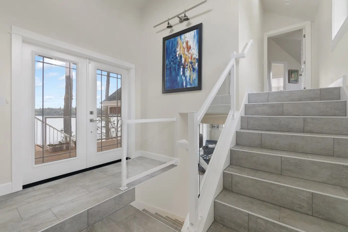 Modern entryway with gray tiled floors and stairs, white walls, glass double doors opening to a lake view, and a colorful abstract painting above the stair railing. Natural light fills the space.