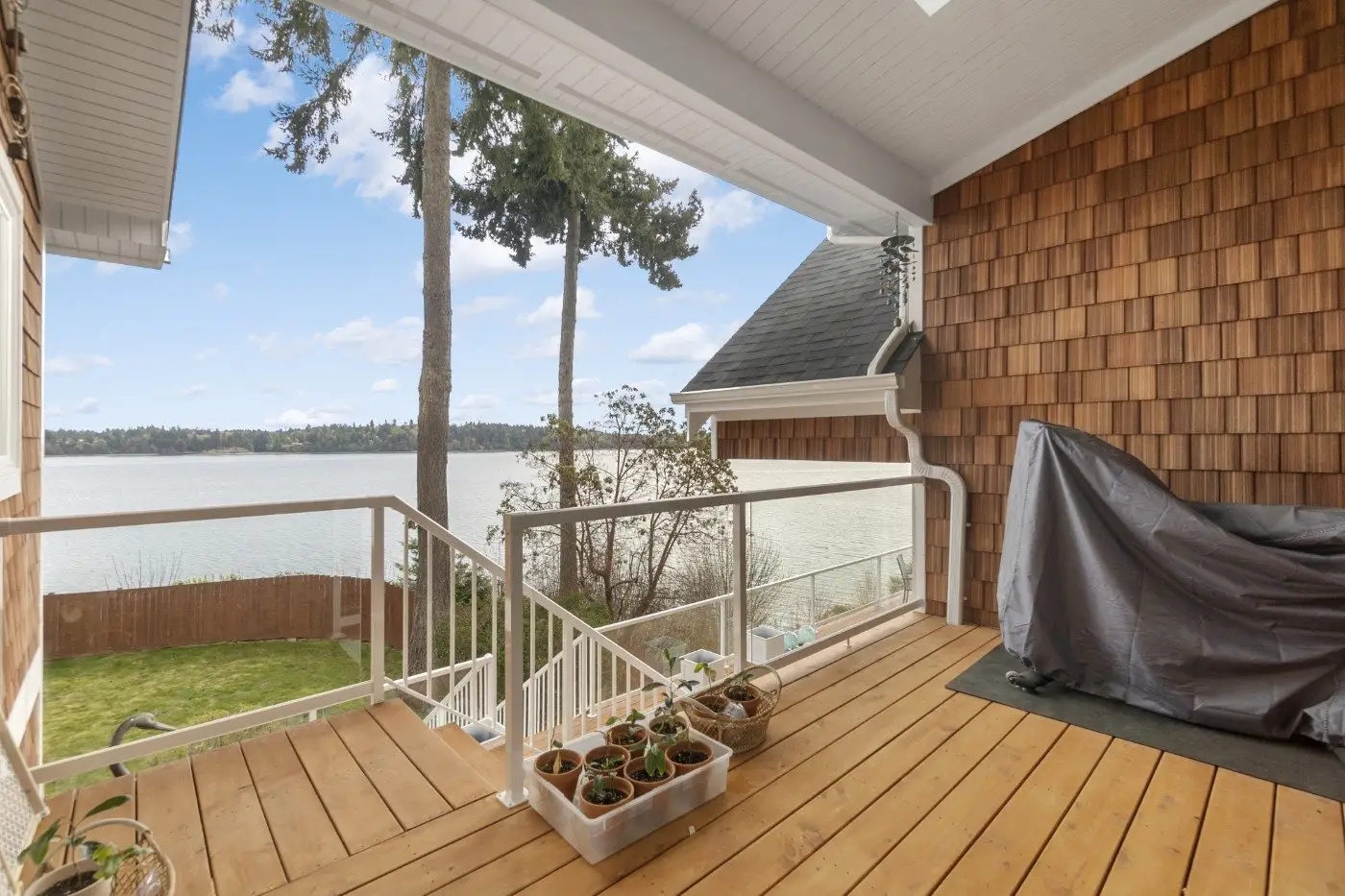 A covered wooden deck with potted plants overlooks a grassy yard and a large lake. Trees and a small building are visible, with clouds and blue sky reflected on the water. A grill is covered on the right side of the deck.