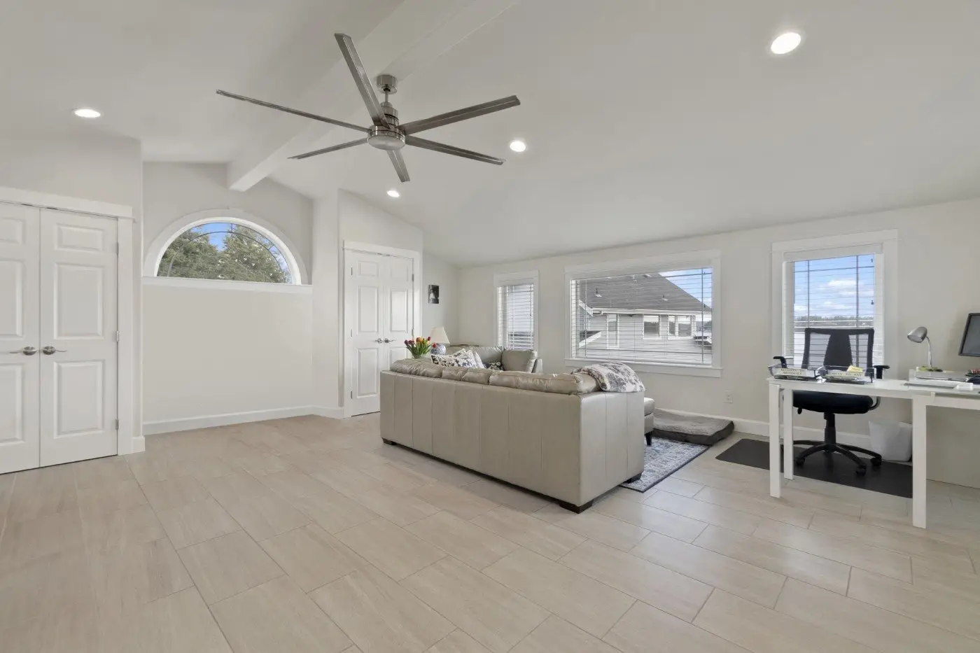 A spacious, modern living room with light wood floors, gray sectional sofa, home office desk, large windows, ceiling fan, and white walls. Natural light fills the room through multiple windows.