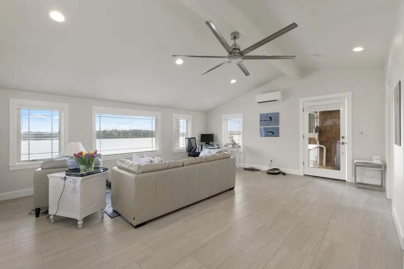 A bright, modern living room with light-colored tile floors, beige sectional sofa, white side table with flowers, a desk by the windows, ceiling fan, and glass door leading to another room. Large windows let in natural light.