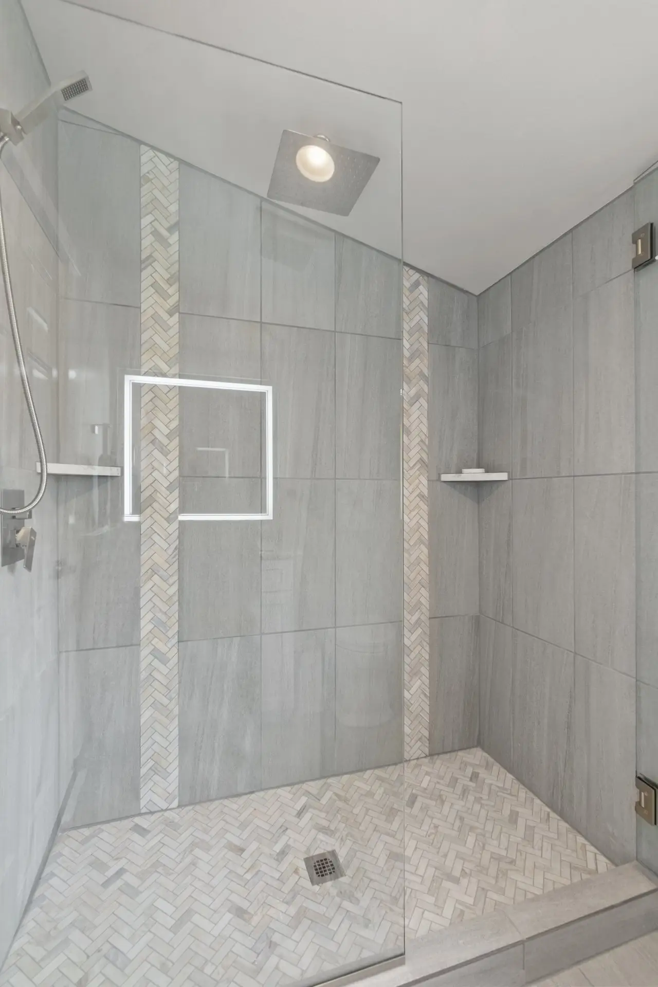 Modern walk-in shower with gray tiled walls and chevron-patterned floor. Vertical mosaic tile accents decorate the wall, and a square rain showerhead is mounted on the ceiling. A lit mirror and corner shelves are visible.