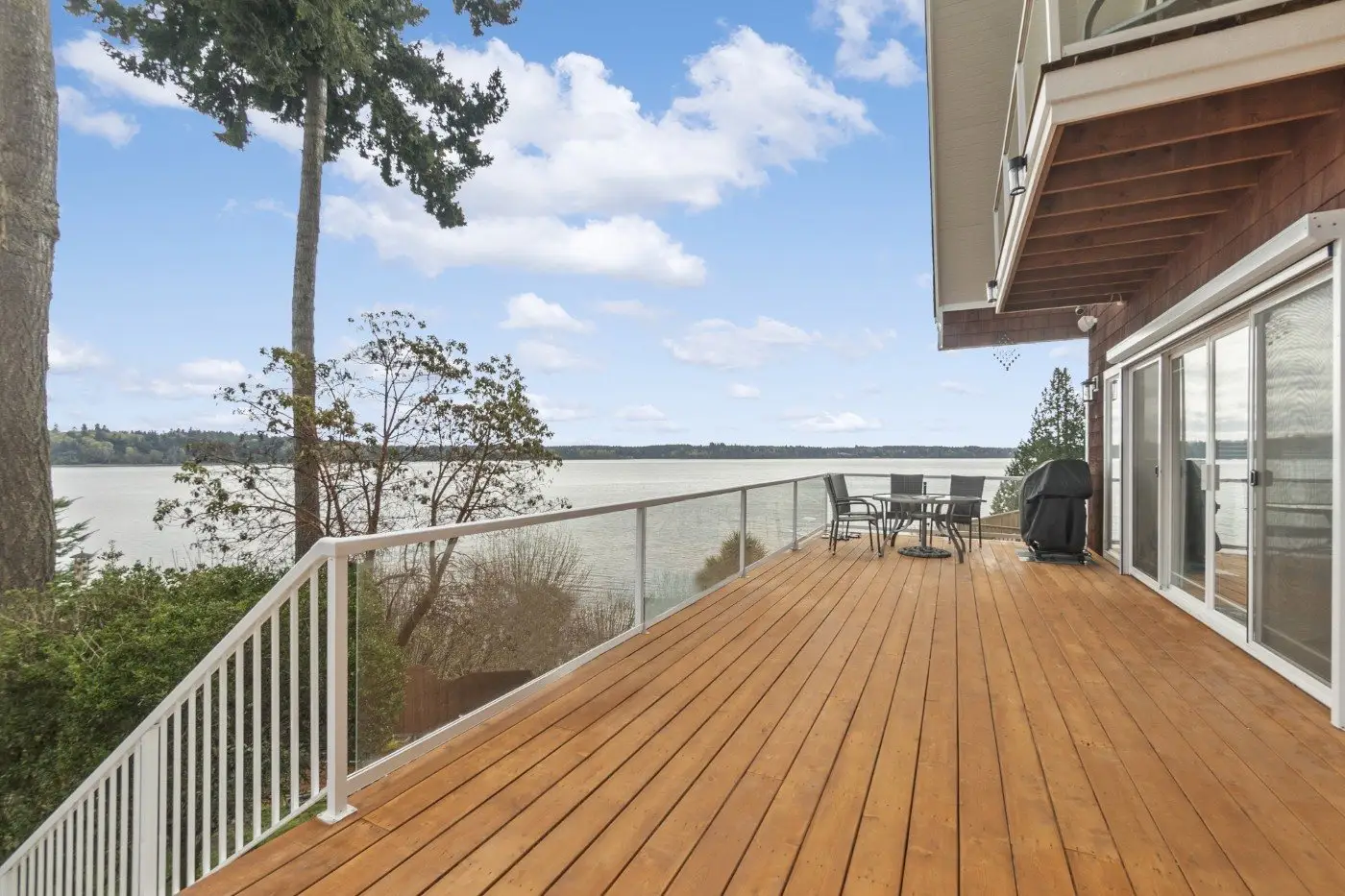 Spacious wooden deck with white railing overlooking a calm lake, surrounded by trees. The deck has a patio table, chairs, and a grill, with a clear blue sky and scattered clouds in the background.