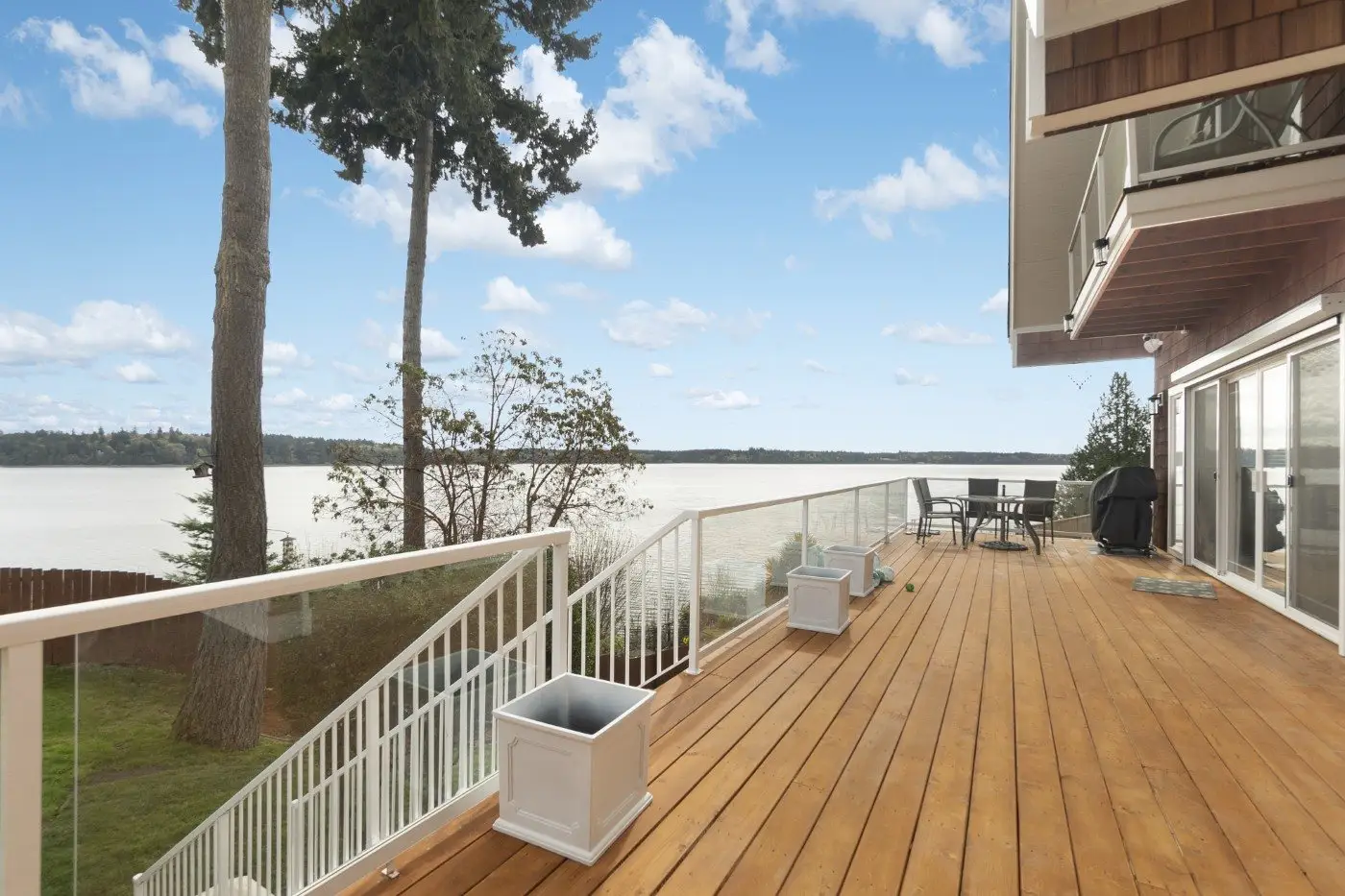 A spacious wooden deck with glass railings overlooks a calm body of water, with trees and a grassy yard nearby. Outdoor furniture and empty planters are arranged on the deck under a blue sky with scattered clouds.