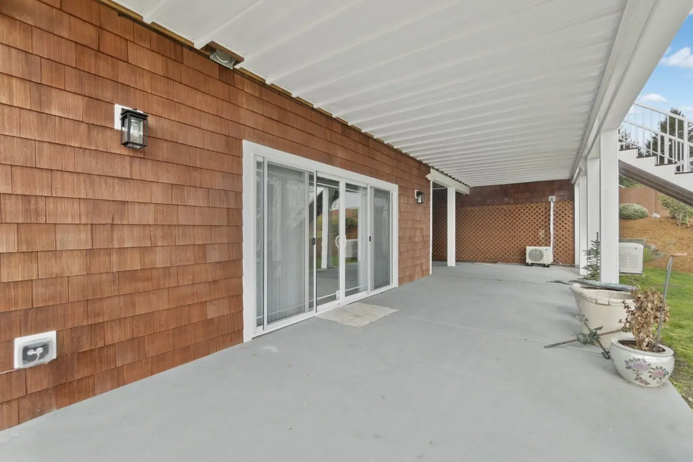 A covered patio with brown shingle siding, sliding glass doors, wall-mounted lights, potted plants, and a concrete floor, overlooking a grassy area with a lattice fence on the right side.