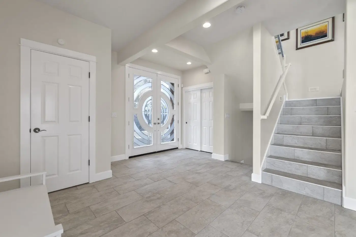 A modern entryway with light gray tile flooring, double front doors with circular glass designs, a staircase with gray steps, and white doors and trim. Walls are light beige with recessed lighting.