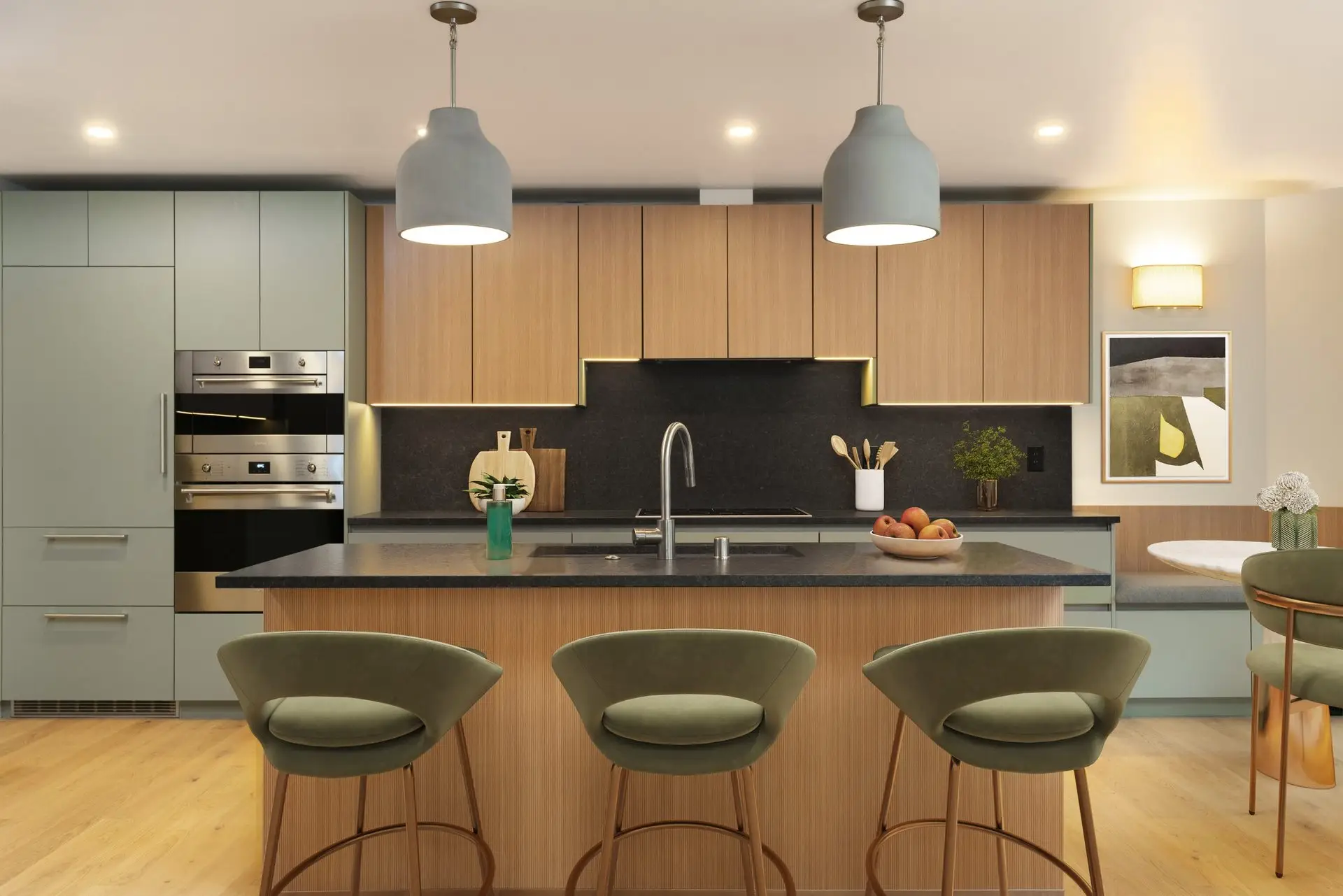 Modern kitchen with light wood cabinets, a central island with a sink, three green velvet bar stools, built-in stainless steel ovens, pendant lights, and minimalist decor on counters and walls.