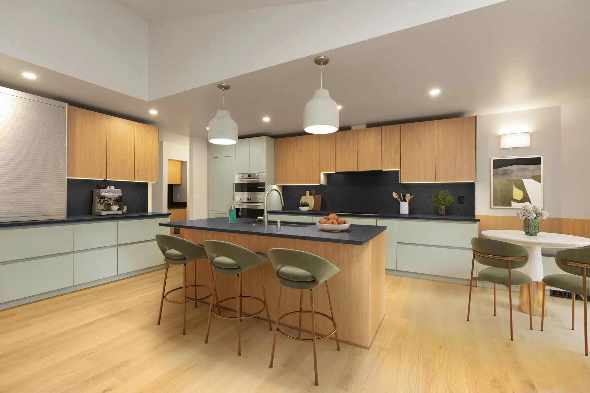 Modern kitchen with light wood floors, an island with four green barstools, pendant lights above, light wood and pale cabinets, built-in appliances, a round dining table with chairs, and wall art.