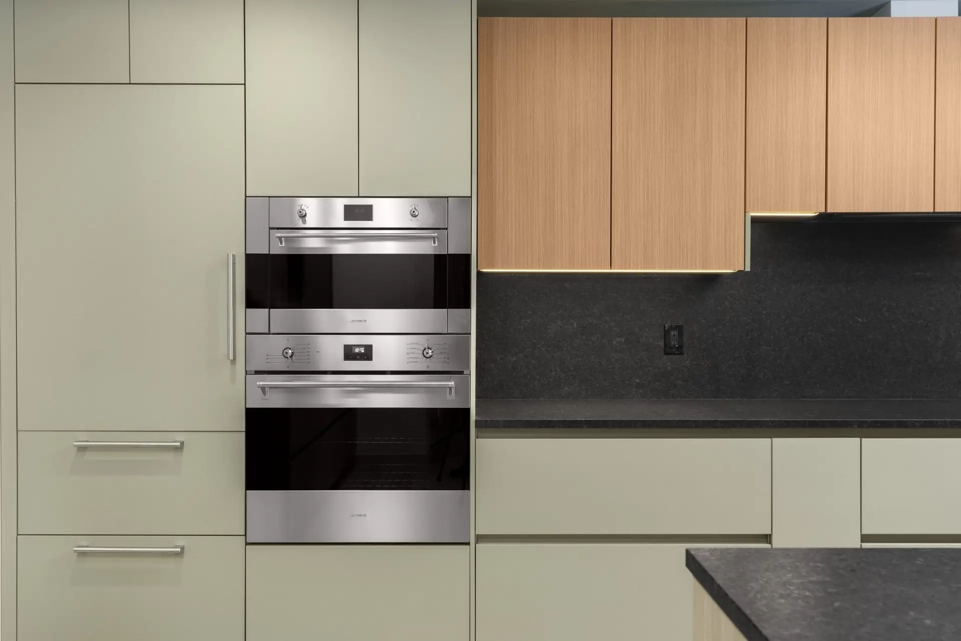 Modern kitchen with light green cabinets, built-in stainless steel double oven, wood upper cabinets, black countertop, and a black backsplash. The design is sleek and minimalistic.