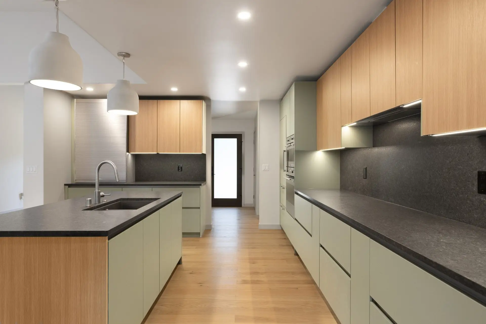 Modern kitchen with light wood and sage green cabinets, black countertops, two pendant lights over an island with a built-in sink, light wood flooring, and recessed ceiling lights.