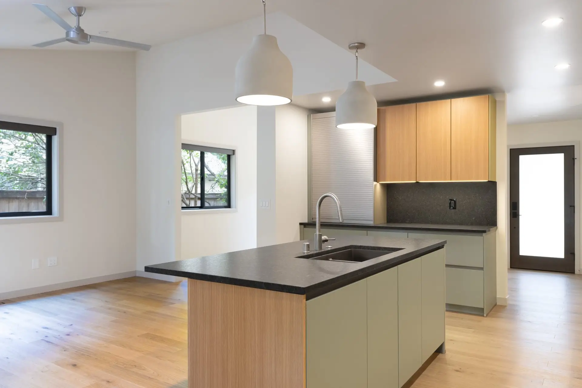 Modern kitchen with light wood floors, pale green cabinets, black countertops, a central island with a sink, pendant lights, and large windows letting in natural light. A door is visible at the back.