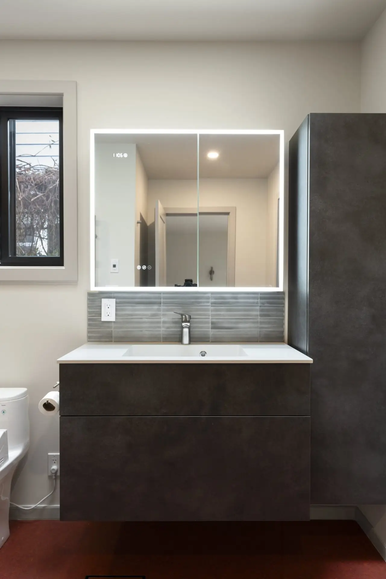 A modern bathroom features a floating dark gray vanity with a white sink, a large backlit mirror, gray tile backsplash, a toilet, and tall cabinet, with neutral walls and a window on the left.