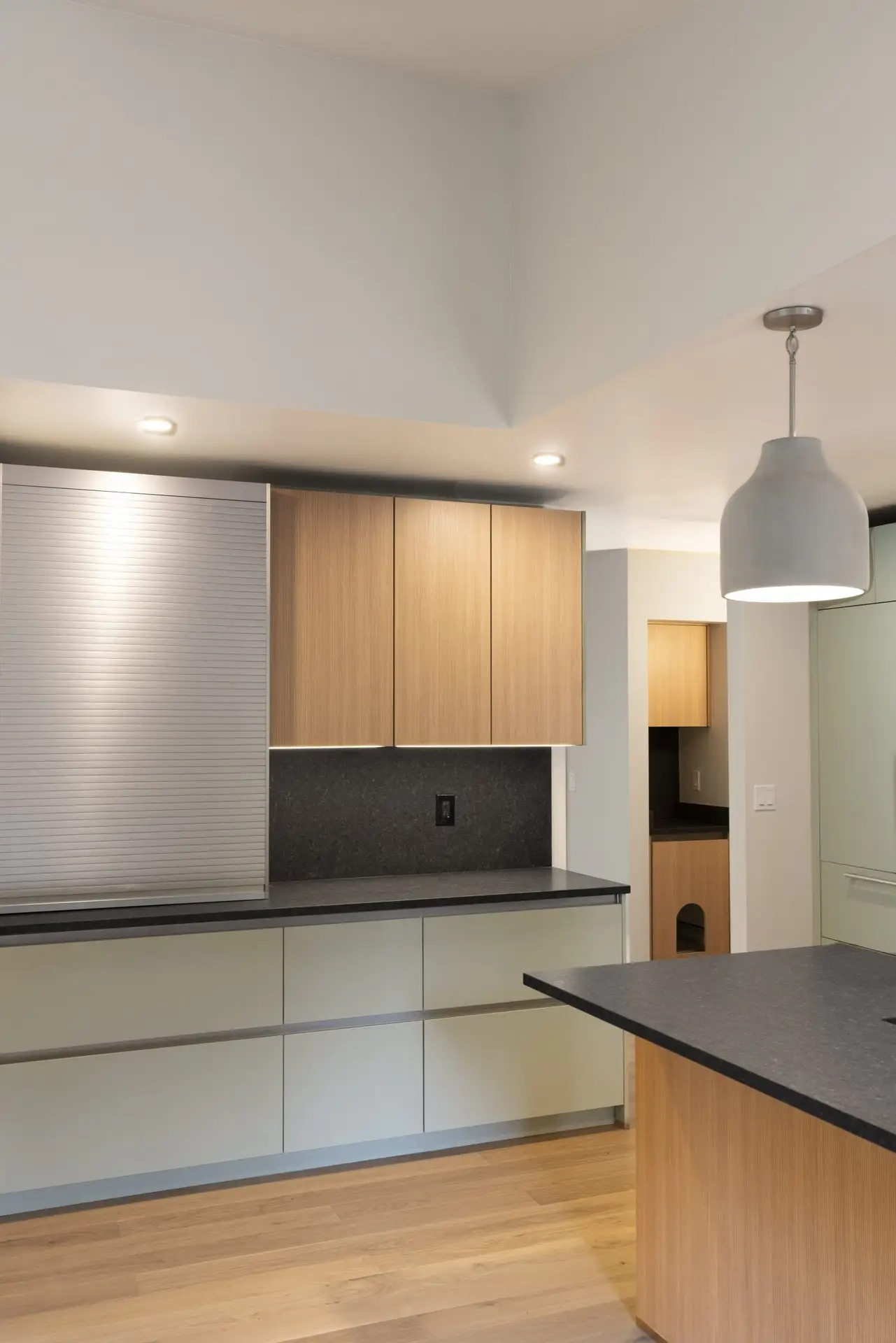 A modern kitchen with light wood cabinets, matte lower drawers, black countertops, recessed lighting, a silver pendant lamp, and light wood flooring. A doorway leads to another room in the background.