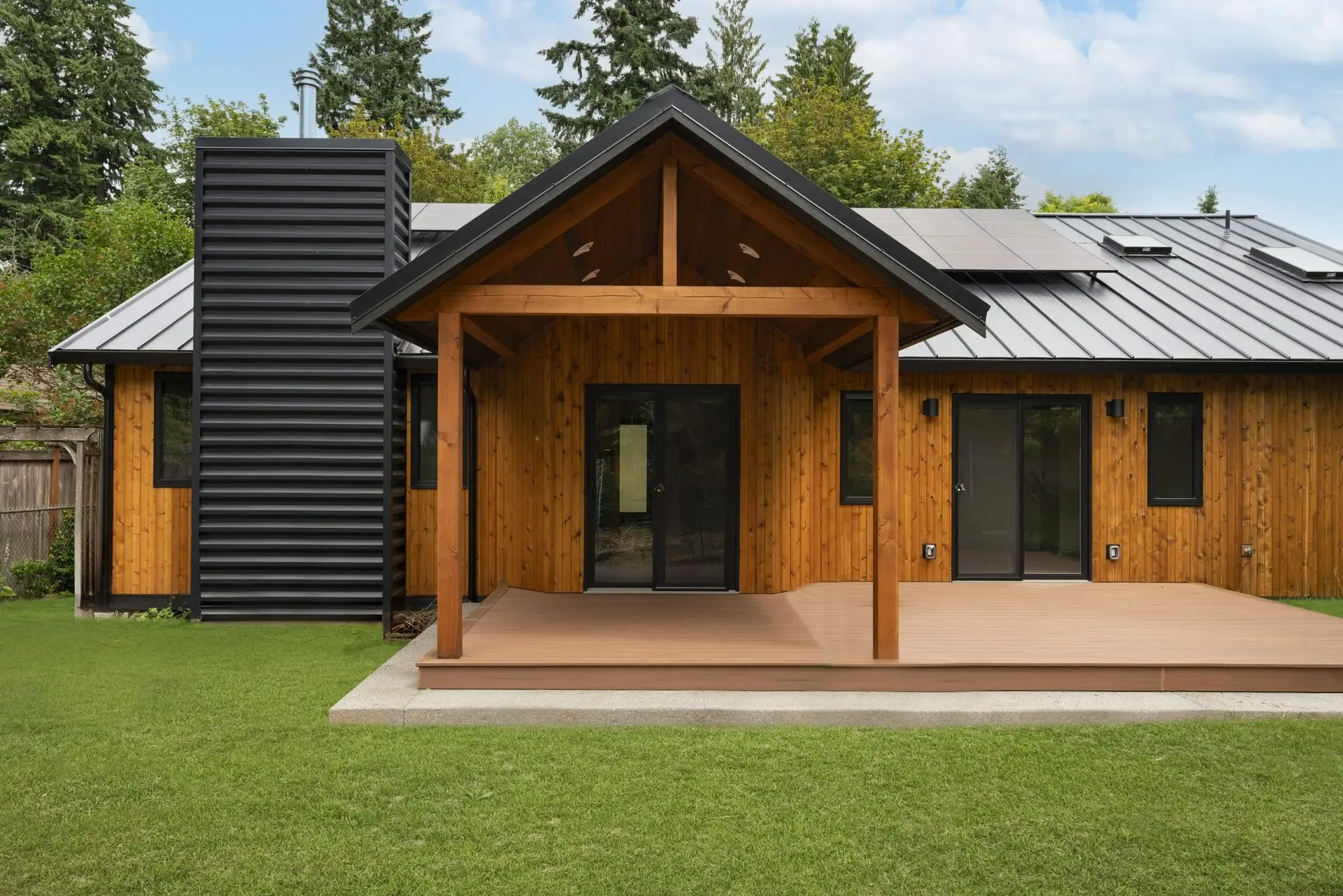 Rear exterior of the Westside Olympia Remodel with cedar siding, black metal roof with solar panels, covered timber-frame porch, and composite deck