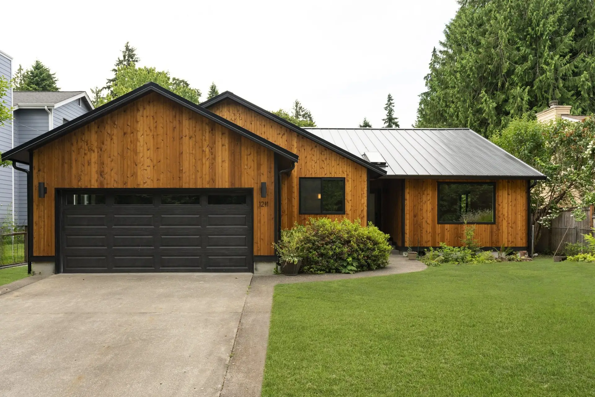 Street view of the Westside Olympia Remodel showing cedar siding, black garage door, standing-seam metal roof, and landscaped front lawn