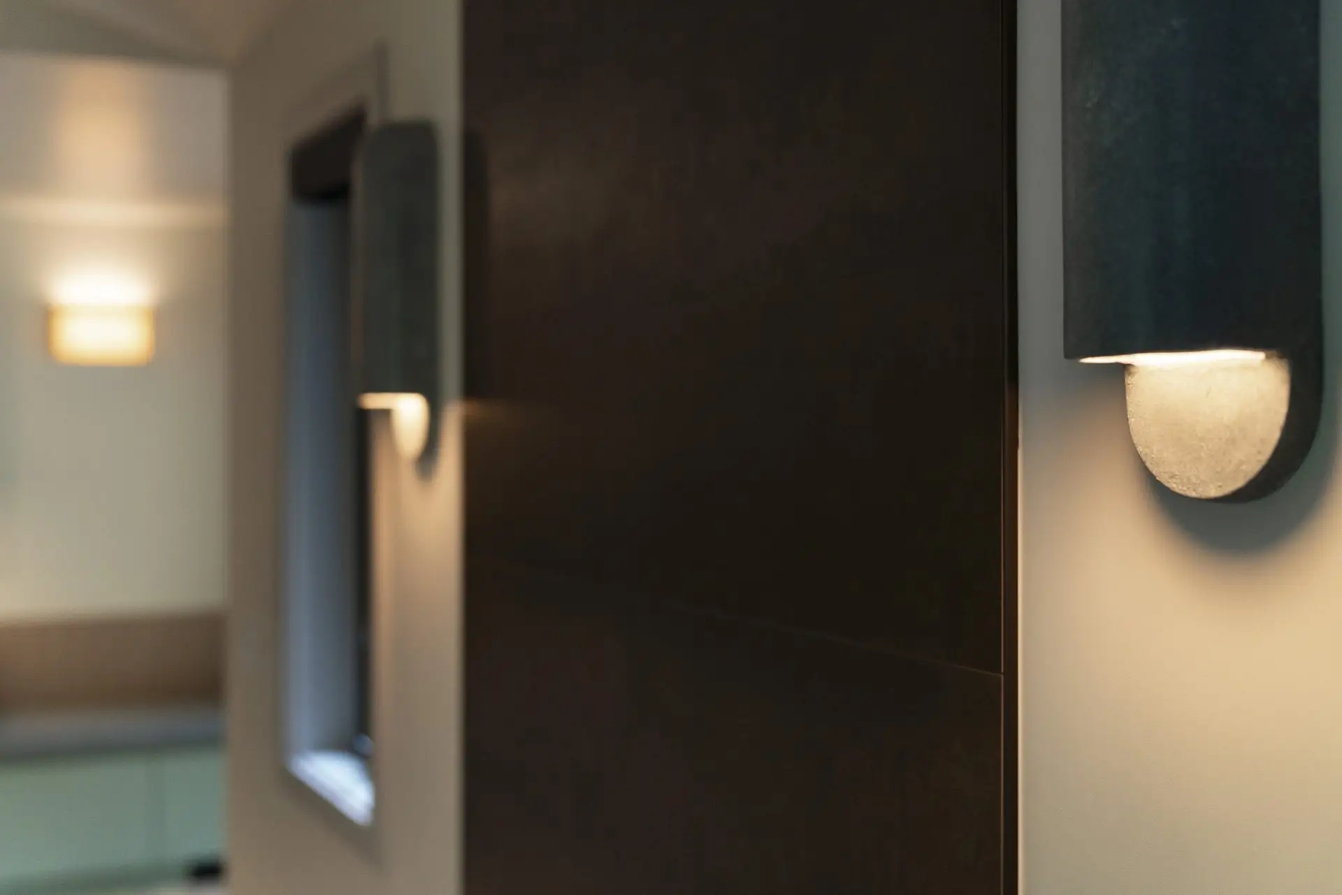 Close-up of a modern wall sconce with a soft, warm light on a beige wall. In the background, there is another sconce and a window, both slightly out of focus, creating a cozy indoor atmosphere.