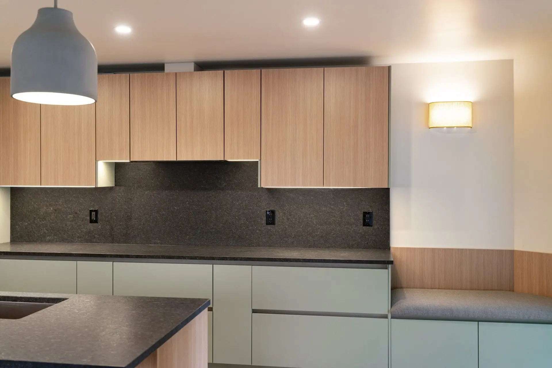 Modern kitchen interior with light wood cabinets, dark countertops, a built-in bench with a gray cushion, pendant and wall lighting, and neutral-toned walls. No appliances or decor are visible.
