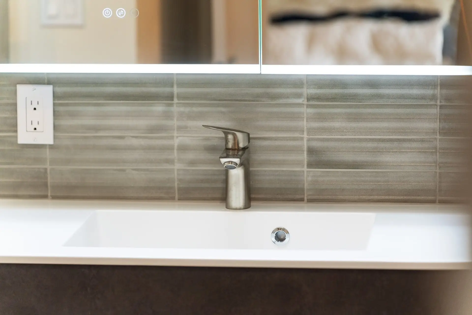 A modern bathroom sink with a sleek silver faucet, set against a wall with horizontal gray tiles and a large mirror above. An electrical outlet is visible to the left of the sink.